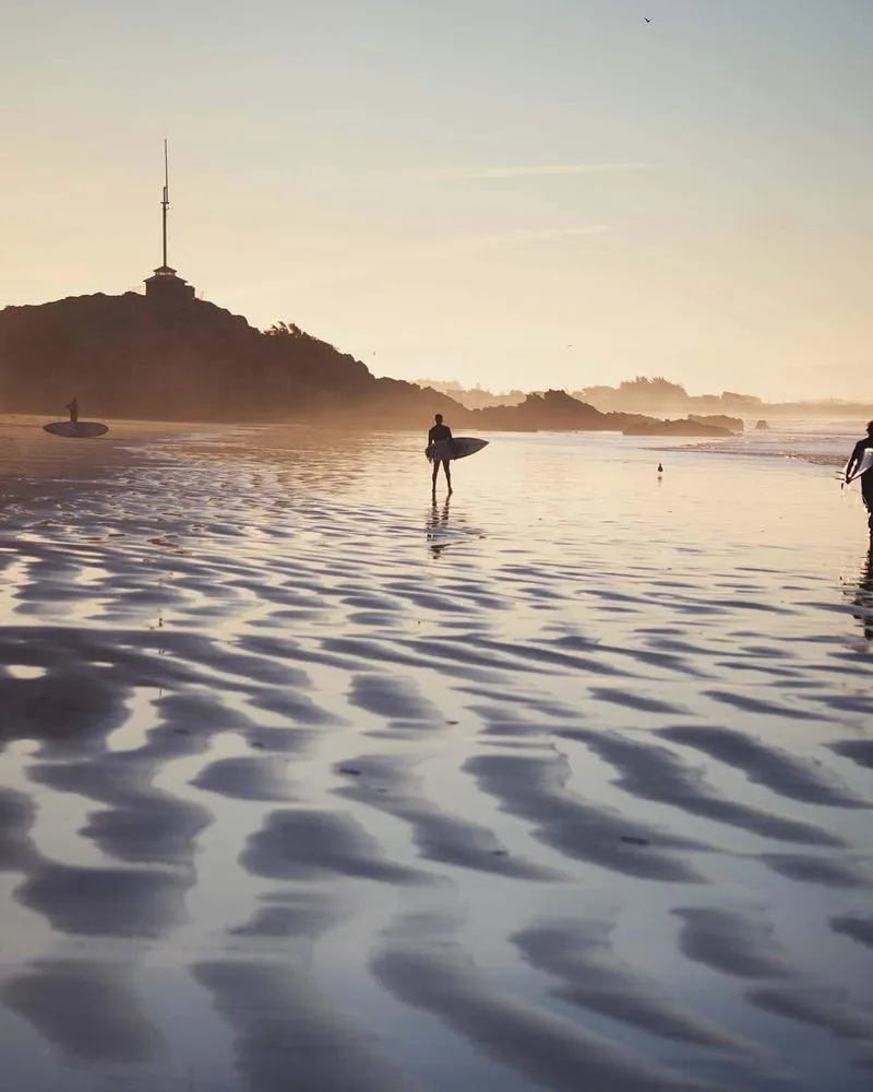 People walking on Scarborough Beach with surfboards, Cave Rock in the background.