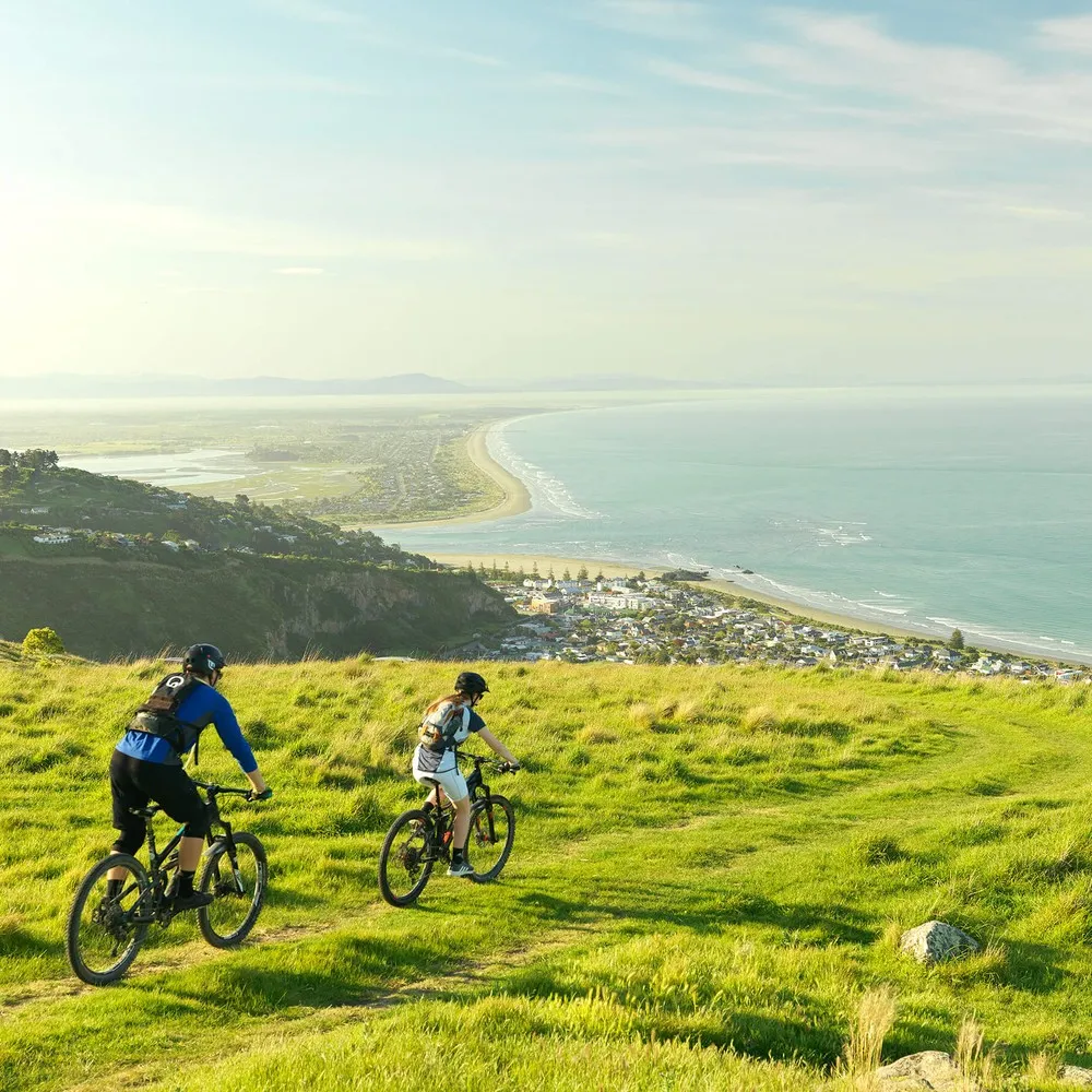 Two mountain bikers on the Port Hills with the Christchurch coastline in the background.