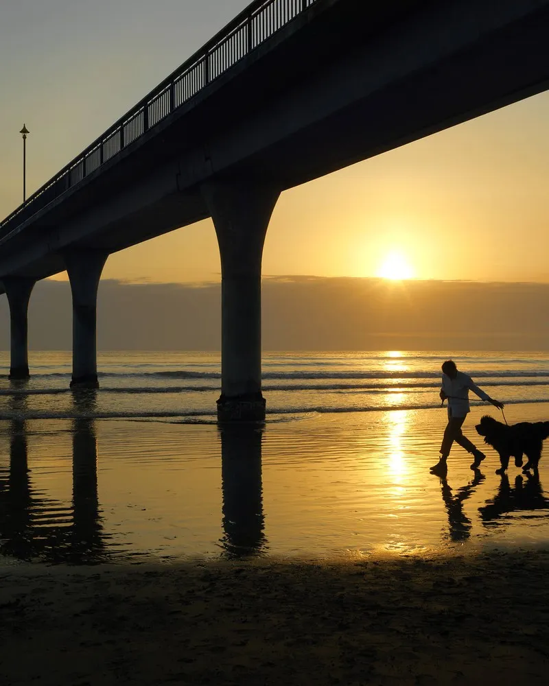 Man walking his dog under the New Brighton Pier with the sunrise in the background.