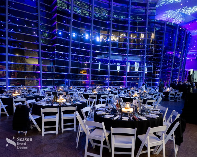 A dinner set up in the atrium area of the Art Gallery. Round tables with black cloths and white chairs. Candles in the centerpieces and fairy lights are strung across the atrium walls