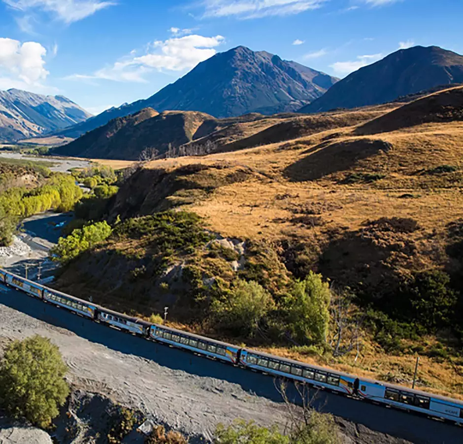 Kiwirail Great Journeys of New Zealand Tranzalpine train travels along the banks of the river with mountains behind 