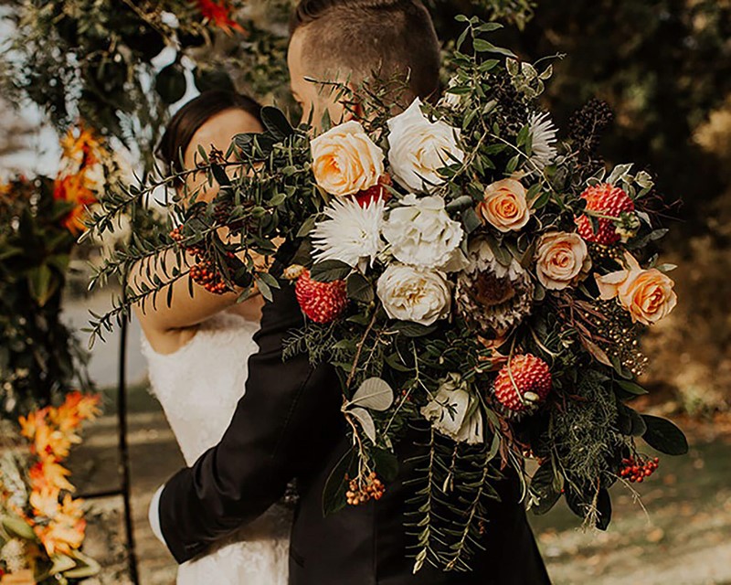 A wedding couple poses for photos, their faces are hidden behind a large autumn theme bouquet 