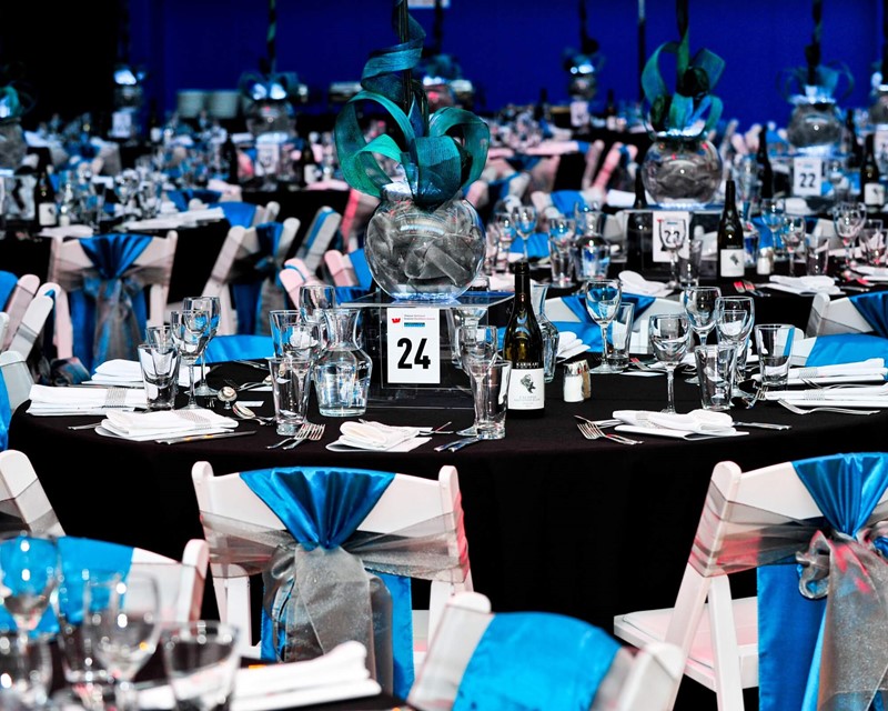 Collective Concepts Business Awards night, round table with black cloth. Blue and silver sashes are tied into bows on the chairs