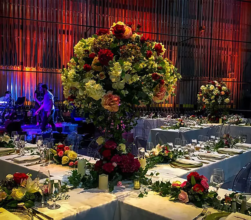 Gala dinner event set up, trestles are set into cross shapes. In the center of the cross are tall striking floral centers. branching out from the center across the length of each table lays greenery with smaller bouquets at intervals 