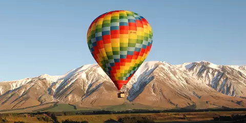 Image shows a colorful hot air balloon in flight across a rural landscape, in the background snow caped mountains. 