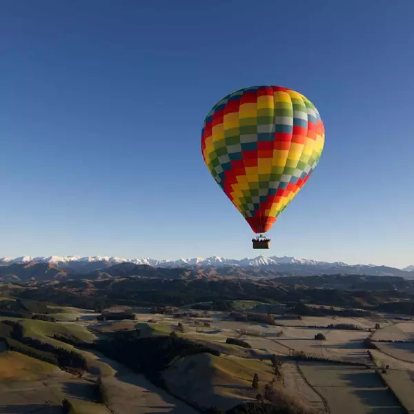 Image shows a colorful hot air balloon in flight across a rural landscape, in the background snow caped mountains. 