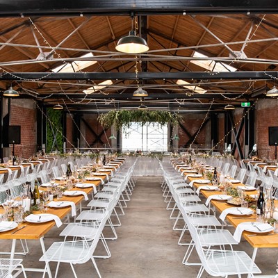 Long tables, timber tops and white chairs set for a function. String fairy lights loop between the exposed beams