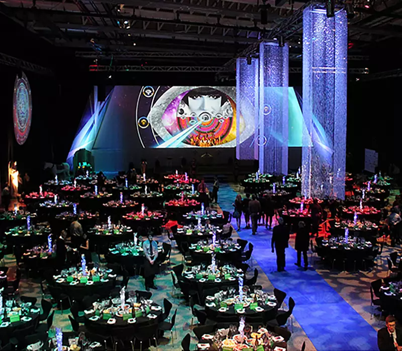 Multi Media Systems gala function set up. Function space is set with round tables, At the front of the room art work image is shown on screen with similar repeated on the sound walls