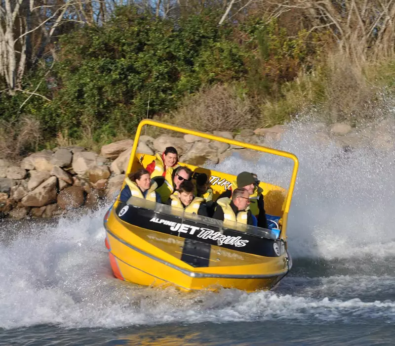 A yellow and orange jet boat is the main focus of the image. The boat is mid spin, water thrown up behind the engine and the guests are wearing yellow lifejackets leaning to one side
