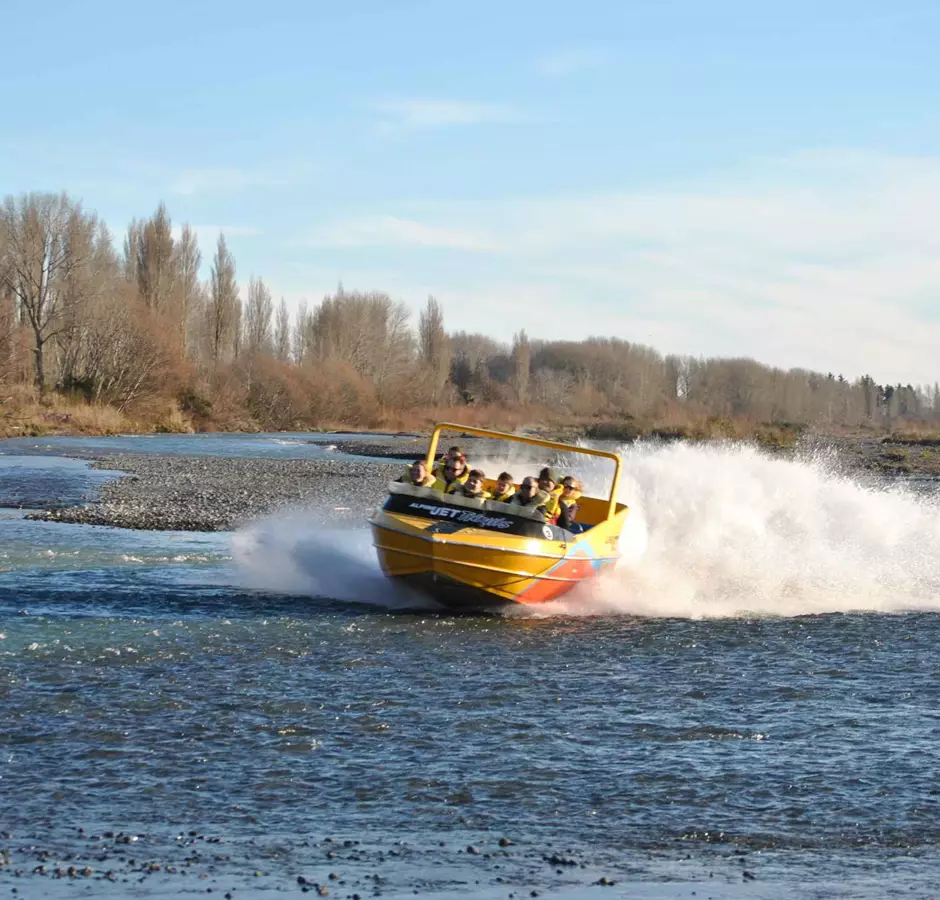 A yellow and orange jet boat heading towards the camera, along the shallow river 